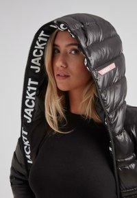 Mujer rubia que lleva una camiseta de canalé negra y una chaqueta acolchada negra con capucha, con el logotipo "JACK1T" en el interior de la capucha y en la etiqueta exterior.