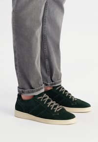 Zielone zamszowe trampki z jasnoszarymi sznurowadłami, białymi gumowymi podeszwami i teksturowanymi akcentami po bokach. Noszone z zrolowanymi szarymi dżinsami.