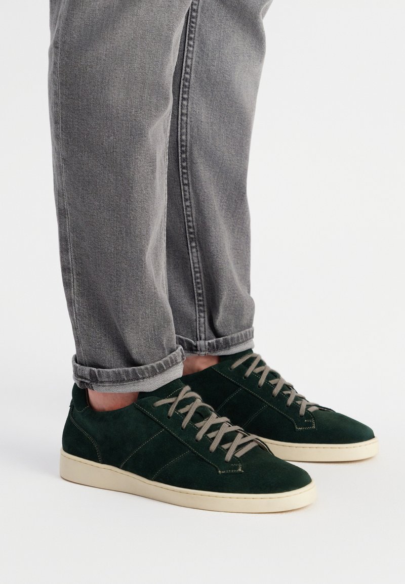 Zielone zamszowe trampki z jasnoszarymi sznurowadłami, białymi gumowymi podeszwami i teksturowanymi akcentami po bokach. Noszone z zrolowanymi szarymi dżinsami.