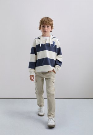 Sudadera a rayas en crema y azul marino, con un bolsillo canguro y capucha con cordón. Pantalones cargo beige con bolsillos laterales y zapatillas blancas.