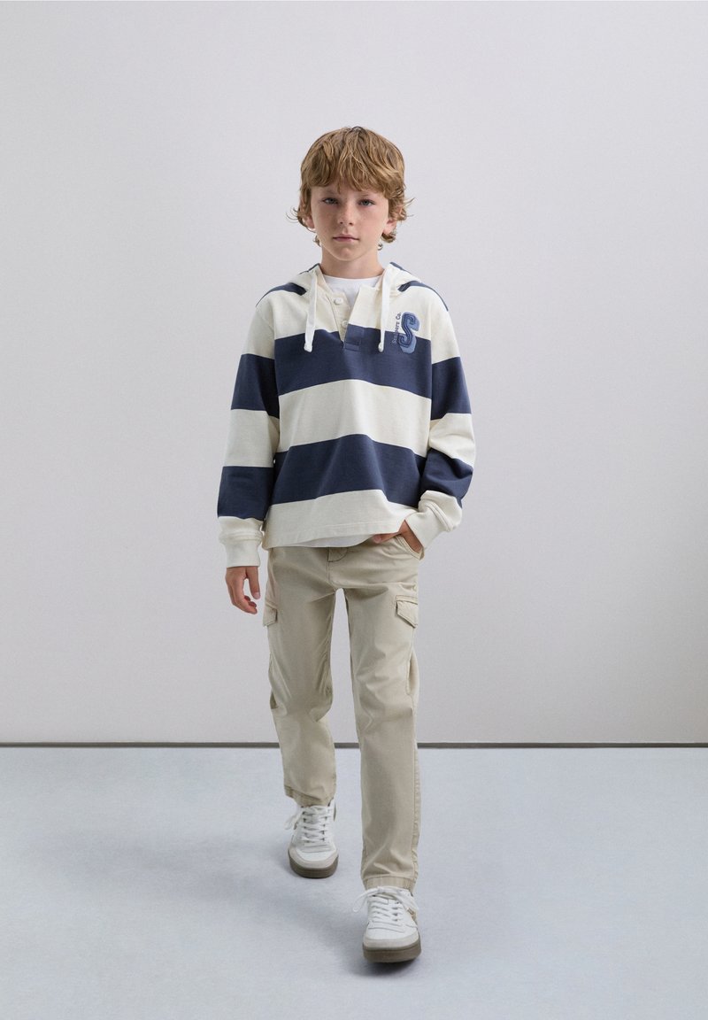 Sudadera a rayas en crema y azul marino, con un bolsillo canguro y capucha con cordón. Pantalones cargo beige con bolsillos laterales y zapatillas blancas.