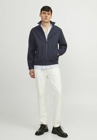 Tumesinine lukuga jakk kõrge kraega, kerge kangas, elastsed mansetid, kantud helesinise särgi ja valgete pükstega, koos mustade kingadega.