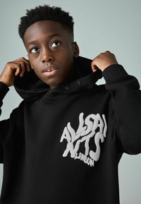 Černá mikina s kapucí, která má velké bílé logo na hrudi, s šňůrkou v kapuci a měkkou texturou. Zobrazena na světlém pozadí.