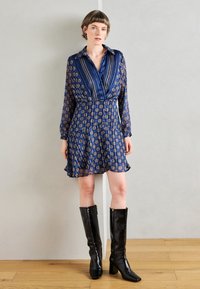 Tamsiai mėlyna suknelė su raštuotu dizainu, ilgi rankovės ir apykakle; derinama su juodais blizgiais aukštakulniais bateliais iki kelių.