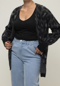 Cardigan à imprimé léopard noir et gris en tissu doux, modèle à ouverture devant, porté sur un top noir avec un jean taille haute bleu clair.