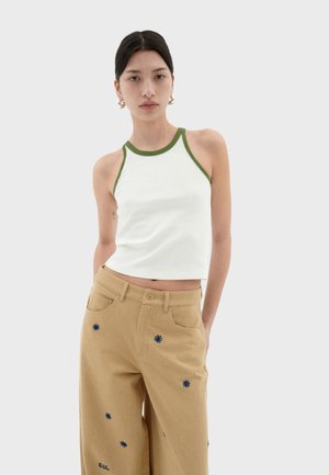 Witte cropped tanktop met groene bies, gecombineerd met lichtbruine broek met blauwe geborduurde bloemenpatronen en zakken.