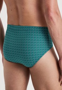 Grüne Schwimmbrieftaschen mit einem geometrischen Muster, das ein wiederholtes griechisches Schlüsselmotiv zeigt. Weicher Stoff, hohe Taille und enganliegende Passform.