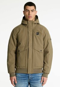 Uomo che indossa una giacca a tema beige con cappuccio, con tasche a risvolto sul davanti e polsini a coste, abbinata a jeans blu, in piedi contro uno sfondo neutro.