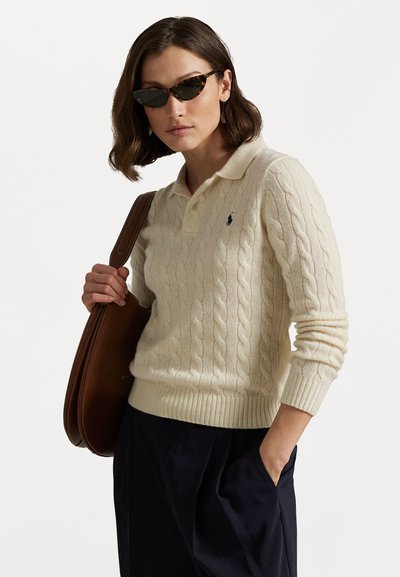 Crème kabelgebreide polo sweater met geribbelde manchetten en zoom, gecombineerd met donkere broek en een bruin leren schoudertas. Donkere zonnebril maakt de look compleet.