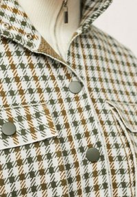 Kurtka w kratę w kolorach zielonym, brązowym i białym. Posiada metalowe guziki na zatrzask, kołnierzyk z zakładką oraz dwie przednie kieszenie. Teksturowana tkanina.