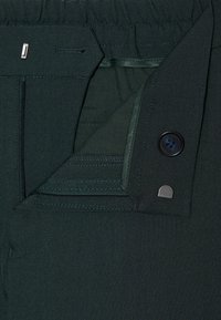 Grüne Stoffhosen mit geradem Schnitt, elastischem Bund, Knopfdetail und subtilen Stickereien. Nahaufnahme des Taschendesigns.