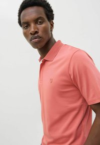 Korall színű pólóing texturált gallérral és diszkrét logóval. Simább anyag, rövid ujjak, testhezálló szabás és klasszikus gallér.