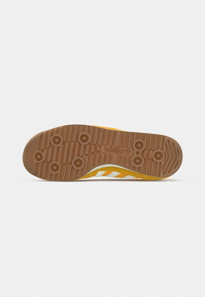 Suela de zapatilla Adidas amarilla con rayas blancas y agarre de goma marrón, que presenta patrones de banda de rodadura circulares y estriados.