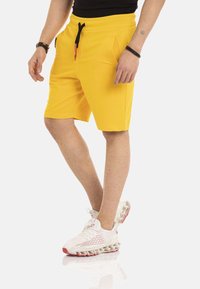Geel sportshort van zacht materiaal, met een elastische tailleband en een zwarte trekkoord, en zijzakken. G gedragen met witte sneakers.
