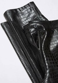 Pantalon noir plié avec une texture brillante de peau de crocodile, avec un bouton et une fermeture éclair visible sur la poche avant.