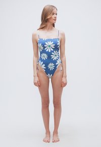 Νεαρή γυναίκα όρθια, ξυπόλυτη, φοράει μπλε ολόσωμο μαγιό με μεγάλα λευκά σχέδια μαργαρίτας και λεπτούς ιμάντες στους ώμους, κοιτάζοντας προς το πλάι.