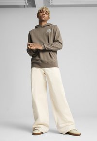 Sudadera marrón con un acabado texturizado, que presenta un logo blanco. Combinada con pantalones de pierna ancha de color crema y zapatillas de color claro.