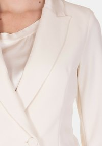 Blazer crema con una texture liscia, rever a lancia e chiusura con un solo bottone. Leggero lucido, vestibilità sagomata e fodera interna a contrasto visibile.