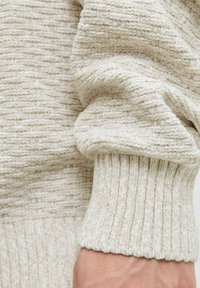 Pull en maille texturée beige clair avec des poignets côtelés, fabriqué dans un tissu doux et tricoté, avec un motif subtil sur l'ensemble du matériau.