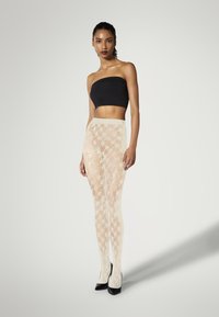 Cremfarbene Strumpfhosen mit Spitzenmuster und durchsichtiger Textur, die ein hellbeiges gewebtes Design aufweisen. Kombiniert mit einem schwarzen trägerlosen Oberteil und schwarzen High Heels.