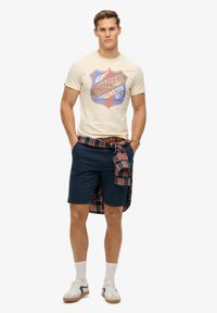 Camiseta beige con un gráfico rojo y azul, pantalones cortos azul marino, camisa de cuadros atada en la cintura, zapatillas blancas y calcetines blancos. Atuendo casual de verano.