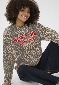 Mikina s leopardím vzorom v béžovej a čiernej farbe s červeným textom "NEW YORK SIMPLICITY". Dlhé rukávy, voľný strih, kombinovaná s tmavými džínami.