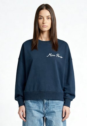 Giovane donna con capelli castani lisci che indossa una felpa blu scuro "Mira Paris" e jeans azzurri, rivolta in avanti su sfondo bianco.