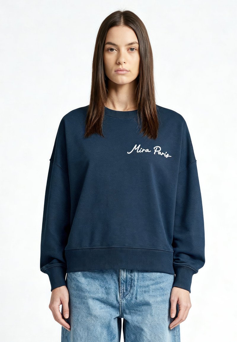 Jeune femme aux cheveux bruns lisses portant un sweat-shirt bleu marine "Mira Paris" et un jean bleu clair, tournée vers l'avant sur un fond blanc.