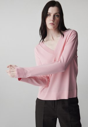 Žena s dlouhými tmavými vlasy, oblečená v světle růžovém svetru s výstřihem do V a černých kalhotách, upravuje svůj levý rukáv pravou rukou.