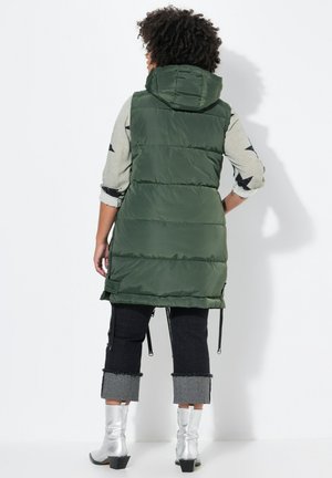 Grüne gepolsterte Weste mit Kapuze, die an den Seiten elastische Zugbänder hat, getragen über einem hellgrauen Langarmshirt mit schwarzen Sternmustern.
