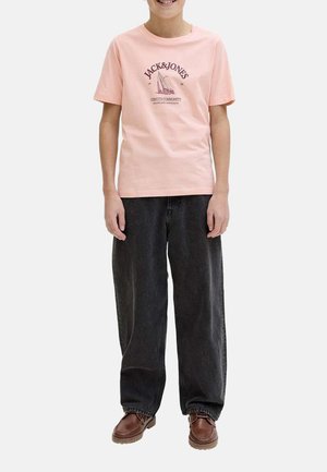 Niño con camiseta rosa claro de Jack & Jones con estampado de velero, jeans negros sueltos y zapatos marrones con cordones, de pie con las manos relajadas.