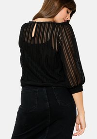 Top nero a maglia con strisce trasparenti e testurizzate; presenta un'apertura a goccia sul retro e maniche a palloncino. Abbinato a jeans neri a vita alta.