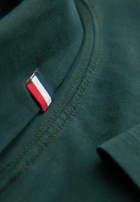 Dunkelgrünes Baumwoll-Polohemd mit einem Kragen, der ein kleines rot-weiß-blaues Etikett trägt. Die Nähte entlang des Randes sind sichtbar.
