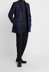 Marineblå ternet frakke med hakket revers, tre knapper og sidelommer, båret over en mørk rullekrave og kombineret med sorte bukser og sko.