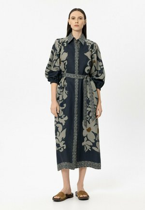 Woman standing wearing a midi dress with large floral patterns, puffed sleeves, a waist tie belt, and brown flat slide sandals.