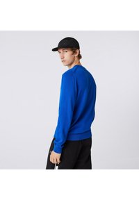 Giovane uomo in un luminoso maglione blu, pantaloni neri e cappellino nero, che guarda indietro oltre la spalla su uno sfondo bianco semplice.