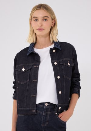 Lyshåret kvinde iført en mørk denimjakke med opkrogede ærmer over en hvid skjorte, med den ene hånd i lommen på hendes matchende mørke denimbukser.
