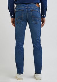 Siniset farkut slim fit -mallissa, joissa on kaksi takataskua ja oranssia ommelta. Täydennetty ruskealla nahkapatchilla vyötäröllä.