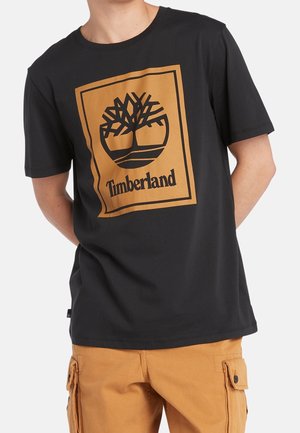 Personne portant un t-shirt Timberland noir avec un grand logo arbre orange et du texte, associé à un pantalon cargo marron clair.