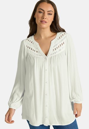 Frau trägt eine weiße Bluse mit langen Ärmeln, Knopfleiste vorne und Spitzenausschnittdetails in der Nähe des Halsausschnitts, kombiniert mit blauen Jeans.