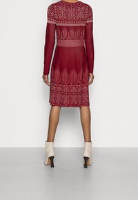 Stickad burgundy klänning med långa ärmar, som har intrikata vita mönster och en figursydd midja. Buren med krämfärgade ankelskor.