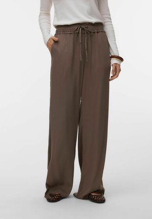Kobieta ubrana w luźne, brązowe spodnie z szerokimi nogawkami ze ściągaczem w pasie oraz brązowe plecione płaskie buty, w połączeniu z białą bluzką z długim rękawem.