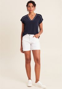 Navy-blaues V-Ausschnitt-Oberteil mit kurzen Ärmeln, kombiniert mit weißen Denim-Shorts. Das Model trägt weiße Sneaker und präsentiert ein lässiges Sommeroutfit.