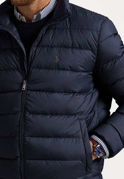 Mand iført en marineblå quiltet jakke over en stribet skjorte og en marineblå sweater, med et blåfaced ur på venstre håndled og hånden i lommen.