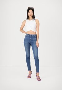 Vit kortad tanktopp med en tvistdetalj, parat med blå skinny jeans och lila remsandaler med klack. Modellen står mot en enfärgad bakgrund.