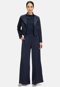 Granatowa zamszowa kurtka o skróconym kroju i wyrazistym kołnierzu, w zestawieniu z szerokimi granatowymi spodniami. Gładka tekstura i minimalistyczne akcenty.