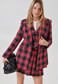 Rot-schwarzes karierter Rockanzug mit doppelreihigem Blazer, körperbetontem Schnitt, strukturiertem Stoff und kontrastierendem weißen Bluse. Detail der schwarzen Handtasche.
