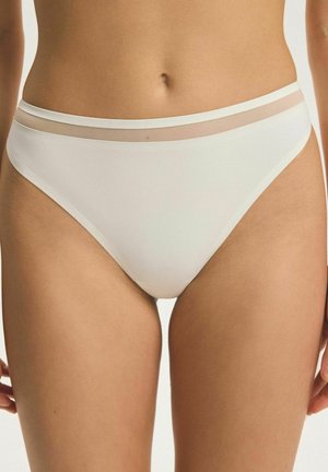 Žena, která má na sobě bílé bikiny s průhledným síťovaným páskem, zobrazena od pasu po horní část stehen na jednoduchém pozadí.