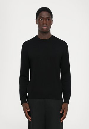 En ung mann står med ansiktet fremover iført en ensfarget svart longsleeve genser og svarte bukser mot en hvit bakgrunn.