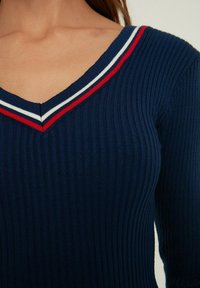 Granatowy sweter prążkowany z dekoltem w kształcie V, z czerwonymi i białymi paskami akcentującymi wzdłuż dekoltu. Gładka tekstura, dopasowany krój.